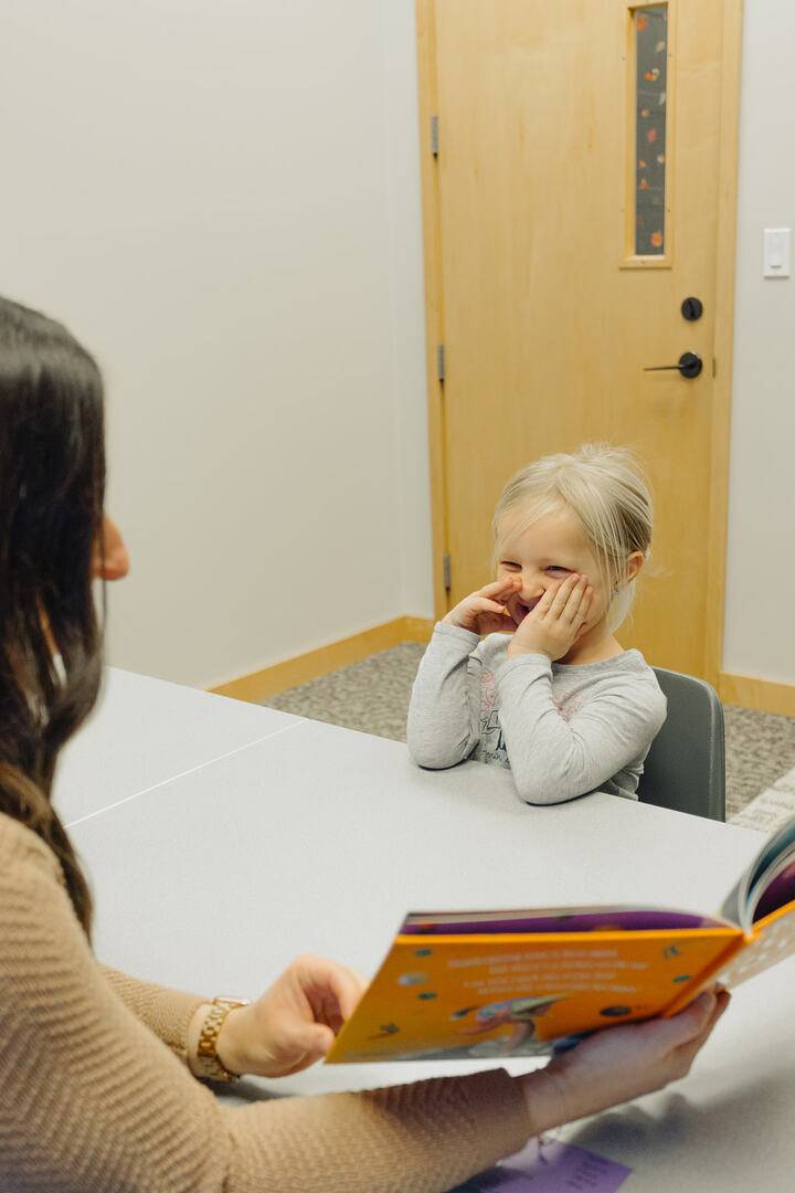 Teacher reading a book to a young smiling girl sitting at a table.