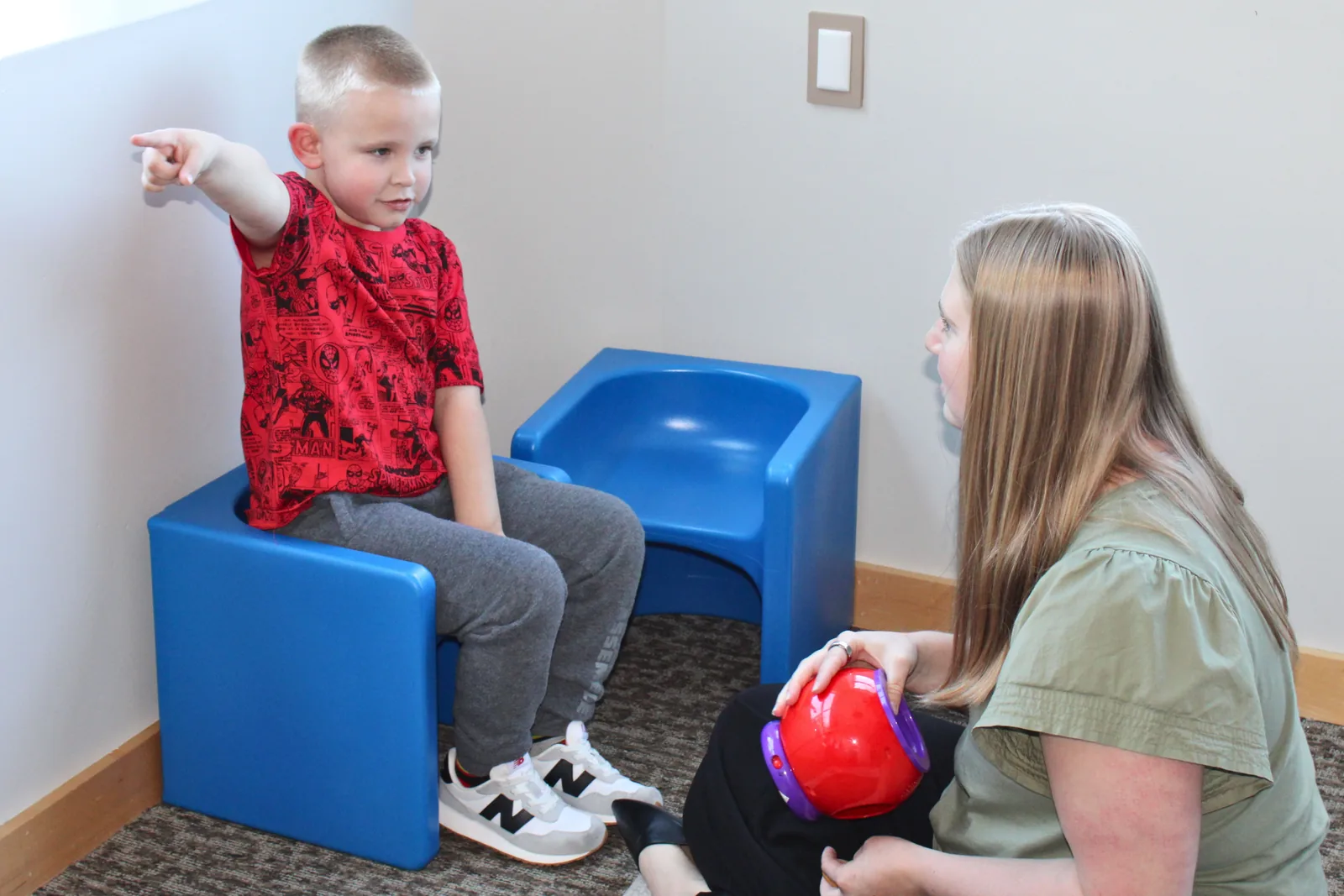 Young boy seated on a cube chair pointing to a feelings choice while a therapist supports him